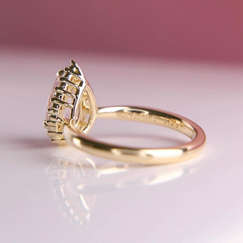 May include: A gold ring with a pear-shaped gemstone. The ring's band and setting are gold. The gemstone is a light pink color and is surrounded by smaller stones. The ring is set against a soft pink background.