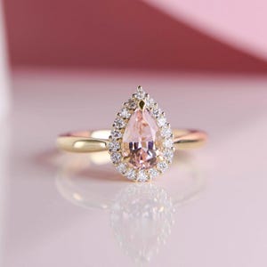 May include: A gold ring featuring a pear-shaped, light pink gemstone surrounded by a halo of small, clear gemstones. The ring's band is a simple, polished gold. The ring is set against a soft pink background.