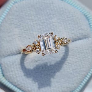 May include: A gold engagement ring with an emerald-cut center stone and smaller accent diamonds. The ring features a delicate floral design on the band, set against a light blue background.