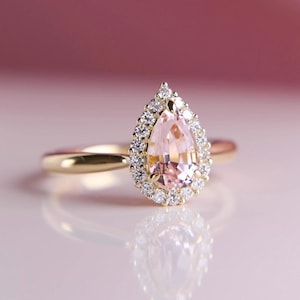 May include: A gold ring featuring a pear-shaped, light pink gemstone surrounded by a halo of smaller, clear stones. The ring's band is a simple, polished gold. The background is a soft, blurred pink.