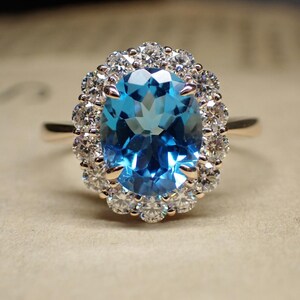 May include: A gold ring featuring a large, oval-cut, light blue gemstone surrounded by a halo of smaller, clear gemstones. The ring's band is a simple, polished gold. The gemstones sparkle, reflecting light.