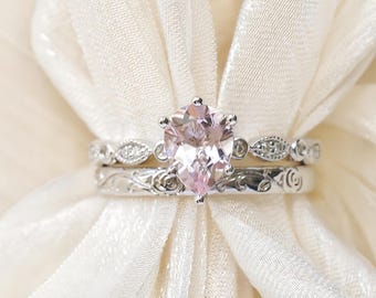 May include: A pair of silver rings. The engagement ring features a pear-shaped, light pink gemstone. The wedding band has a delicate design with small clear stones and a floral pattern. The rings are set against a soft, cream-colored fabric.