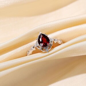 May include: A silver ring with a pear-shaped, deep red gemstone as the focal point. The gemstone is surrounded by a decorative setting with smaller, clear stones. The ring rests on a cream-colored fabric.