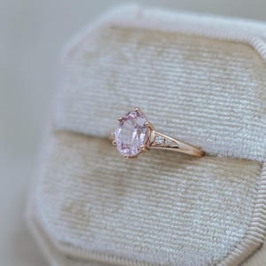 May include: A rose gold ring featuring a large, oval-cut pink gemstone as the centerpiece. The band is adorned with smaller, clear accent stones. The ring is set against a soft, neutral-toned background, highlighting its elegant design.