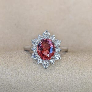 May include: A silver ring featuring a large, oval-cut, pink gemstone surrounded by a halo of smaller, round, white gemstones. The ring's design is a classic halo style, with the central stone standing out.