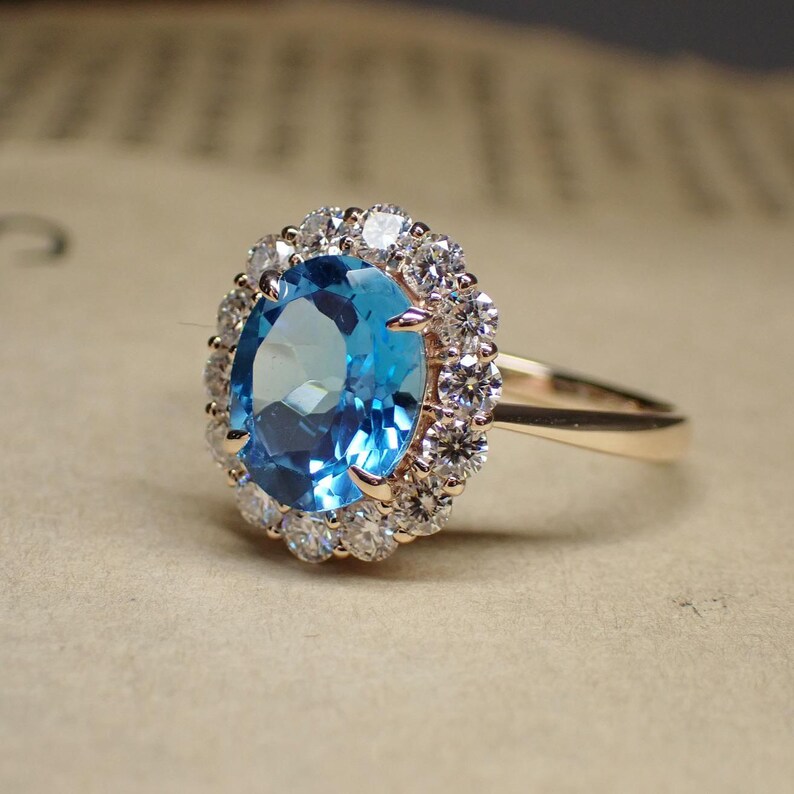 May include: A gold ring featuring a large, oval-cut, light blue gemstone surrounded by a halo of smaller, clear gemstones. The ring's band is a polished gold color, and the setting highlights the central stone.