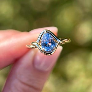 May include: A gold ring featuring a pear-shaped, light blue gemstone. The gemstone is set in a unique, swirling gold band. The ring is a piece of jewelry, likely an engagement or fashion ring.