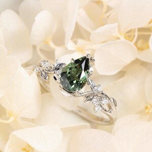 May include: A silver ring featuring a pear-shaped, deep green gemstone as the centerpiece. The ring is adorned with delicate floral and leaf accents, and small, clear gemstones. The ring is set against a backdrop of white flowers.