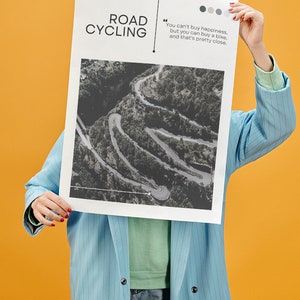 Puede incluir: Una persona sostiene un p&oacute;ster con una foto en blanco y negro de un camino sinuoso en una zona monta&ntilde;osa. El p&oacute;ster tiene el texto "ROAD CYCLING" y una cita: "No se puede comprar la felicidad, pero se puede comprar una bicicleta, y eso es bastante cerca."