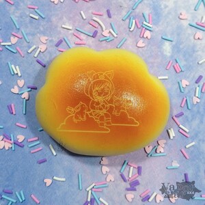 May include: A yellow, cloud-shaped squishy toy with a cartoon illustration of a girl and two animals. The illustration is etched into the surface. The background is a light blue surface scattered with colorful sprinkles and heart-shaped confetti.