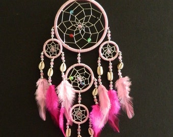 May include: Pink dream catcher with multiple layers of beads, feathers, and woven circles. The dream catcher has a pink and white color scheme.