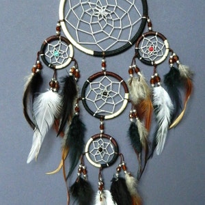 May include: A handcrafted dreamcatcher with a large central hoop and multiple smaller hoops below. The dreamcatcher features a white web design, brown and black accents, beads, and feathers in shades of black, brown, and white. The background is a solid gray.