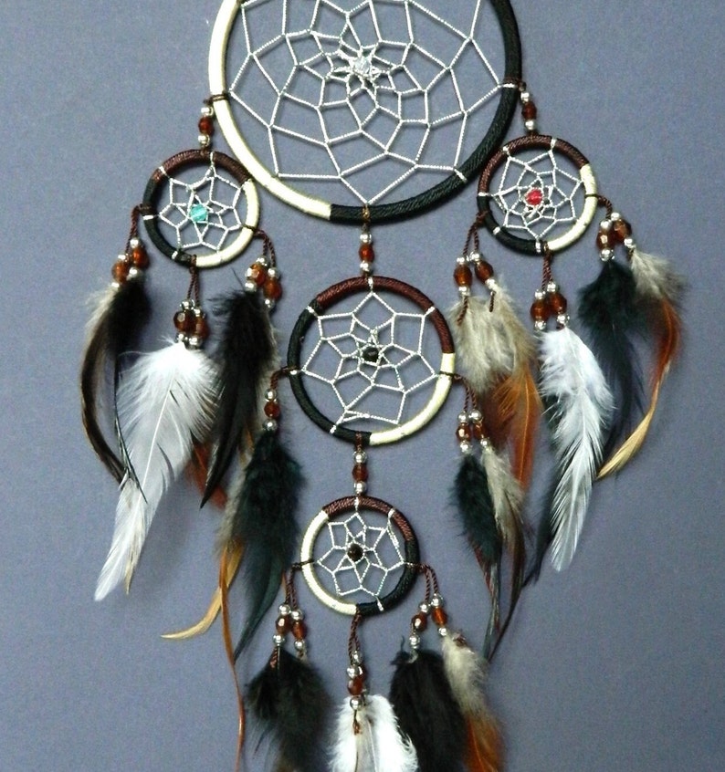 May include: A handcrafted dreamcatcher with a large central hoop and multiple smaller hoops below. The dreamcatcher features a white web design, brown and black accents, beads, and feathers in shades of black, brown, and white. The background is a solid gray.