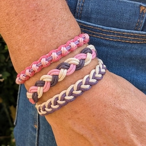 May include: Three braided bracelets in shades of pink, purple, and cream are displayed on a wrist. The bracelets have a woven design, with the colours alternating in each row. The background includes a person's arm and blue denim jeans.