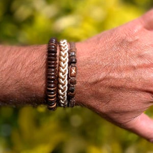 May include: Four bracelets are shown, each with a unique design. The first is made of dark brown wooden beads. The second features a braided pattern with brown and cream strands. The third is a brown bracelet with a wooden bead with a white cross.