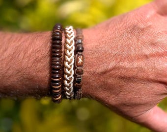 May include: Four bracelets of varying designs and colors are displayed. The first bracelet is made of dark brown wooden beads. The second is a braided design with alternating brown and cream-colored strands. The third is a brown bracelet with a wooden bead with a white cross.