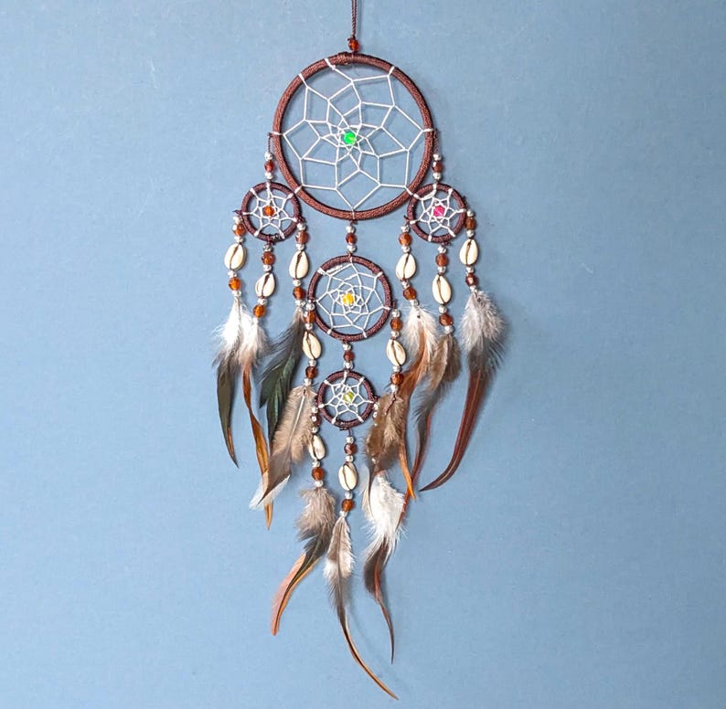 Brown dreamcatcher with 5 rings. The top ring is 9cm wide. All rings have silver webs with a central bead. Four rings hang below the top ring, strung with beads, cowrie shells and black feathers. Length 30cm