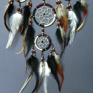 May include: A handcrafted dreamcatcher with multiple circular frames, each featuring a web-like design. The dreamcatcher is adorned with brown, black, and white feathers, along with small beads. The color palette includes shades of brown, black, white, and cream.