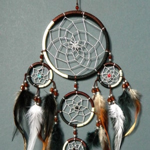 May include: A handcrafted dreamcatcher with a large central hoop and three smaller hoops below. The hoops are adorned with intricate web-like designs, beads, and feathers in shades of brown, black, and white. The dreamcatcher hangs against a gray background.