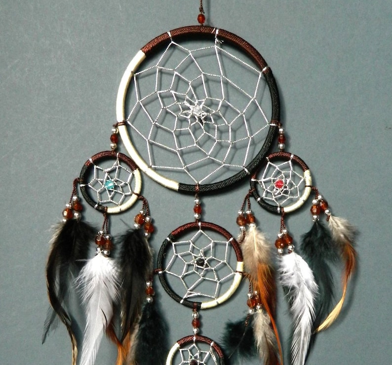 May include: A handcrafted dreamcatcher with a large central hoop and three smaller hoops below. The hoops are adorned with intricate web-like designs, beads, and feathers in shades of brown, black, and white. The dreamcatcher hangs against a gray background.