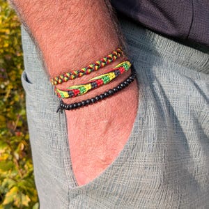 May include: Three bracelets on a wrist. The top bracelet is braided with red, yellow, and green threads. The middle bracelet is woven with the same colors. The bottom bracelet is made of black beads. The person is wearing a light blue shirt.