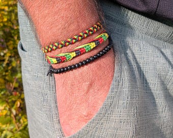 May include: Three bracelets on a wrist. The top bracelet is braided with red, yellow, and green threads. The middle bracelet is woven with the same colors. The bottom bracelet is made of black beads. The person is wearing a light blue shirt.