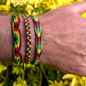 May include: Three colourful woven bracelets in red, yellow, green, and black are displayed on a person's wrist. The bracelets have a braided and woven design. The background is blurred with yellow flowers.
