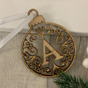 May include: A wooden Christmas bauble ornament with the letter "A" at its centre. The bauble has a detailed, openwork pattern and is hung by a white ribbon. A sprig of evergreen is visible in the bottom right corner of the image.
