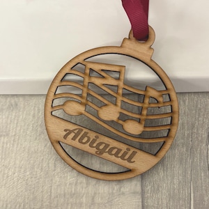 May include: Wooden Christmas bauble with a musical note design and the name "Abigail" engraved. The bauble is round with a dark red ribbon for hanging. The design features a treble clef and musical notes.