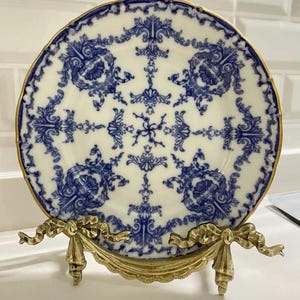 Peut inclure: Une assiette en porcelaine bleue et blanche avec un motif floral d&eacute;coratif, pr&eacute;sent&eacute;e sur un support en m&eacute;tal dor&eacute;.