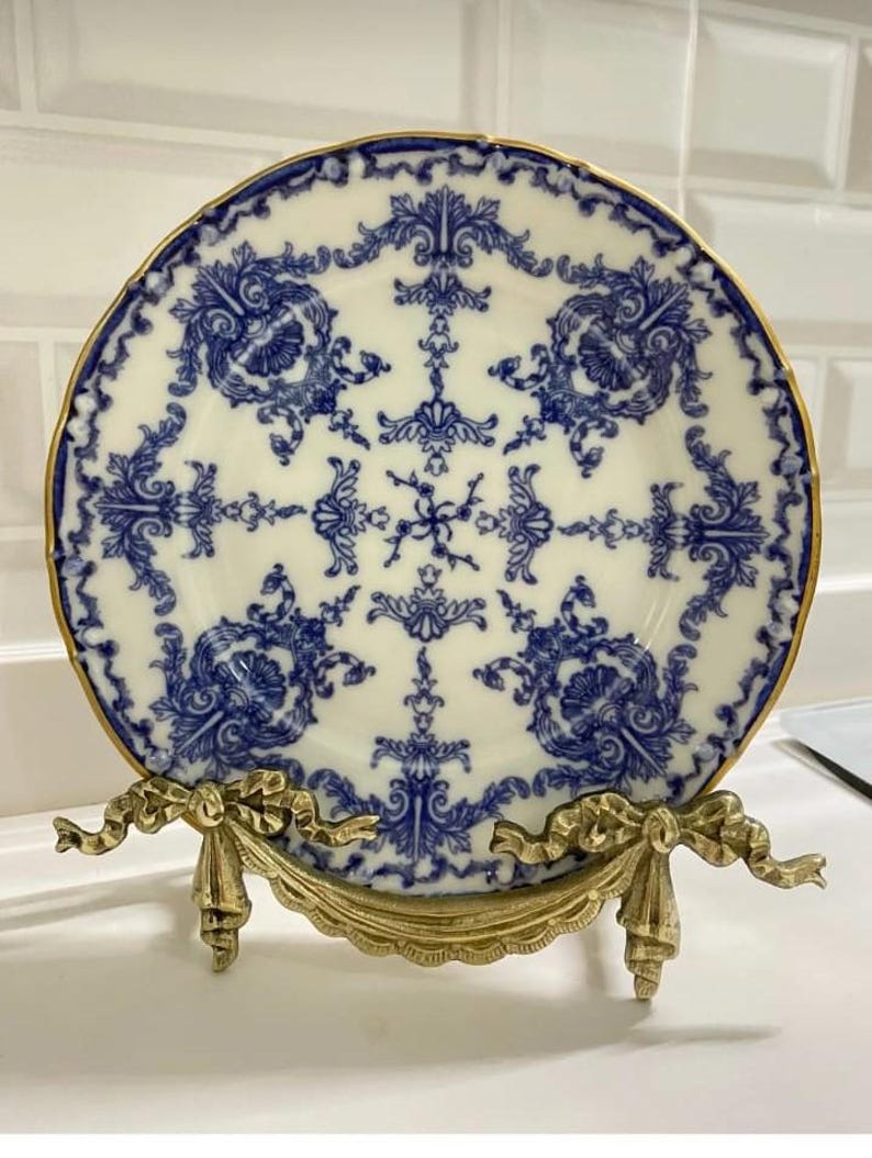 Peut inclure: Une assiette en porcelaine bleue et blanche avec un motif floral d&eacute;coratif, pr&eacute;sent&eacute;e sur un support en m&eacute;tal dor&eacute;.