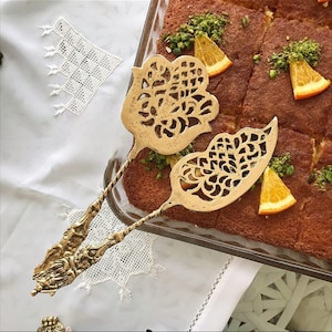 May include: Two ornate gold-colored serving utensils with intricate floral designs. The utensils are placed near a rectangular dish of baked goods garnished with orange slices and green sprinkles. The background includes a white tablecloth with lace details.