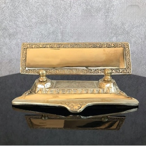 Puede incluir: Un tintero de latón vintage con dos tinteros y un portaplumas. El tintero tiene un diseño ornamentado con un borde elevado y detalles intrincados.