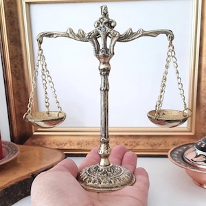 Könnte beinhalten: Eine dekorative Messingwaage mit zwei kleinen Schalen, die an Ketten hängen. Die Waage wird in einer Hand gehalten, was ihr aufwendiges Design und ihre Vintage-Ästhetik zeigt. Der Hintergrund umfasst ein gerahmtes Bild und andere dekorative Gegenstände.