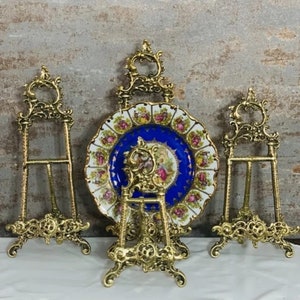 Può includere: Quattro supporti per piatti in metallo dorato decorato con un piatto in porcellana decorativo al centro. Il piatto ha un centro blu con un motivo floreale e accenti dorati.