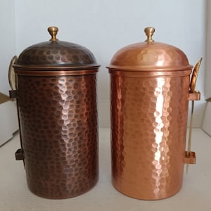 Könnte beinhalten: Zwei Kupferkanister mit gehämmerten Oberflächen und Deckeln. Jeder Kanister hat einen kleinen Löffel an der Seite.
