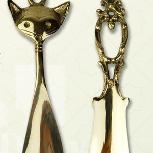 May include: Two decorative brass shoehorns. One is shaped like a cat's head, the other has an ornate handle. Both are polished to a high shine, reflecting the light. The shoehorns are designed for easy use and add a touch of elegance.