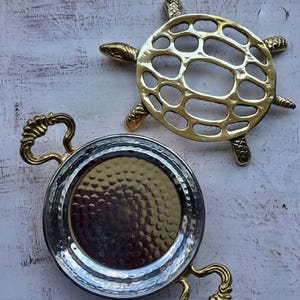 Könnte beinhalten: Zwei dekorative Metallgegenstände: ein goldfarbener Untersetzer in Schildkrötenform und ein silberfarbenes Tablett mit goldenen Griffen. Das Tablett hat eine gehämmerten Textur auf der Innenseite. Der Schildkröten-Untersetzer ist aus goldfarbenem Metall mit einem Ausschnittmuster.