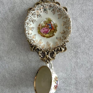 Könnte beinhalten: Ein Vintage-Tassen- und Untertassen-Set mit einer verzierten Wandhalterung aus goldfarbenem Metall. Die weiße Porzellantasse und Untertasse haben einen goldenen Rand und Blumen- und Paarillustrationen. Die Wandhalterung hat ein dekoratives, gerolltes Design.
