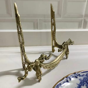 Peut inclure: Un support d&eacute;coratif en m&eacute;tal dor&eacute; avec un design orn&eacute; de n&oelig;uds et de rubans. Le support est con&ccedil;u pour tenir une assiette ou un autre objet d&eacute;coratif.