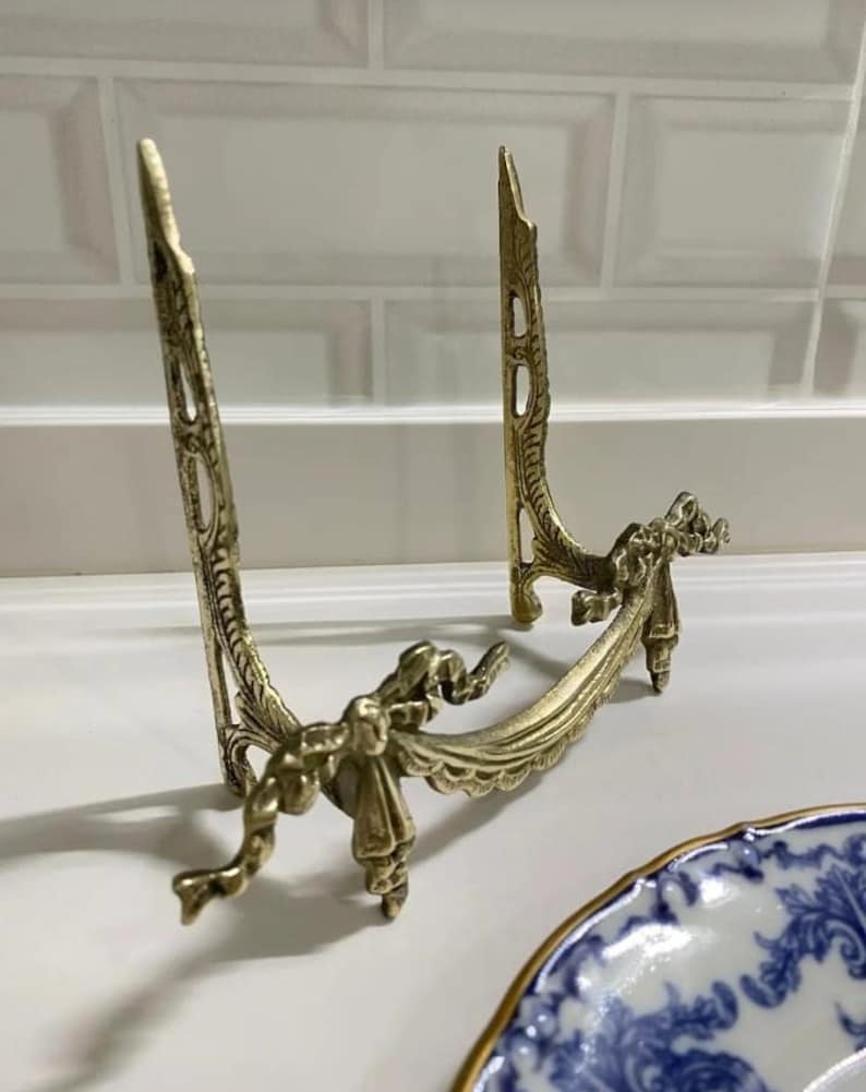 Peut inclure: Un support d&eacute;coratif en m&eacute;tal dor&eacute; avec un design orn&eacute; de n&oelig;uds et de rubans. Le support est con&ccedil;u pour tenir une assiette ou un autre objet d&eacute;coratif.