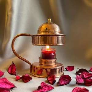 Könnte beinhalten: Kupferner Räucherofen mit einer Kuppel und einem Griff. Ein kleines Glas mit einer roten Kerze steht auf einer unteren Plattform. Der Räucherofen ist von roten Rosenblättern umgeben.