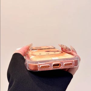 May include: A clear phone case with a glittery, orange-toned border. The case features a cartoon cat design on the back. The phone case is held by a person wearing a black long-sleeve shirt.