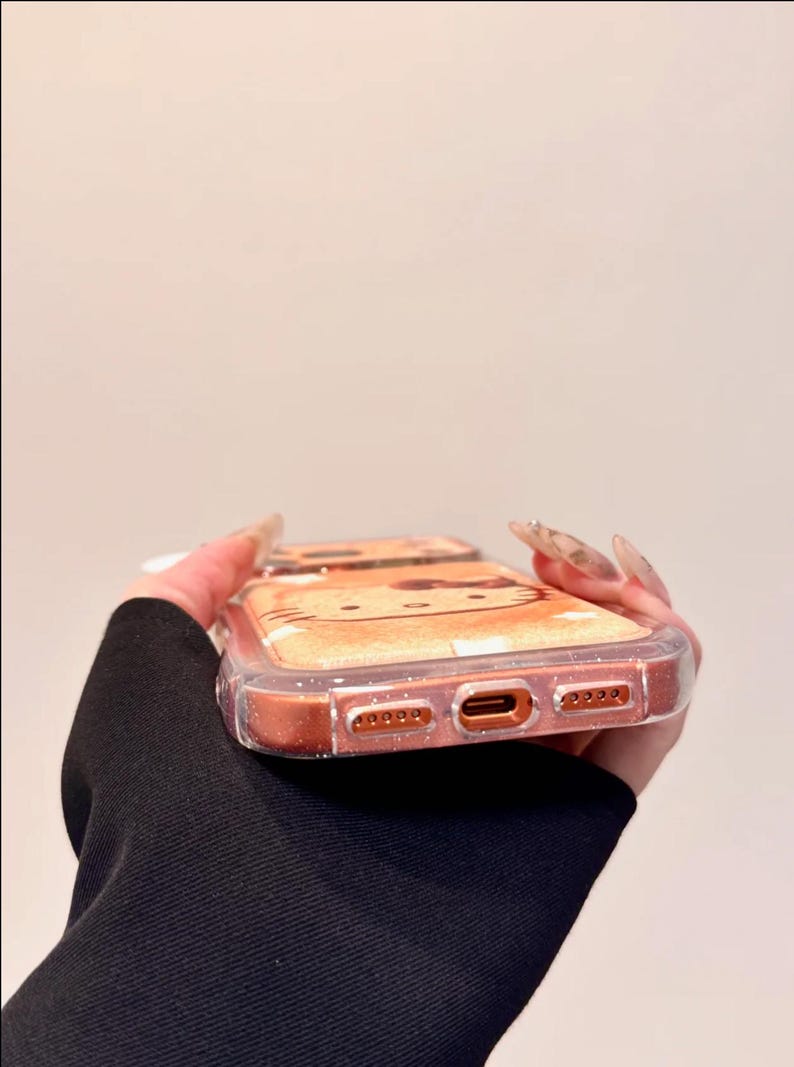May include: A clear phone case with a glittery, orange-toned border. The case features a cartoon cat design on the back. The phone case is held by a person wearing a black long-sleeve shirt.