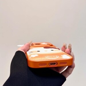 May include: Orange phone case with a Hello Kitty design. The case has a glittery finish and a cartoon image of Hello Kitty. It has cutouts for the phone's camera, charging port, and speakers. The text "Hello Kitty" is visible.