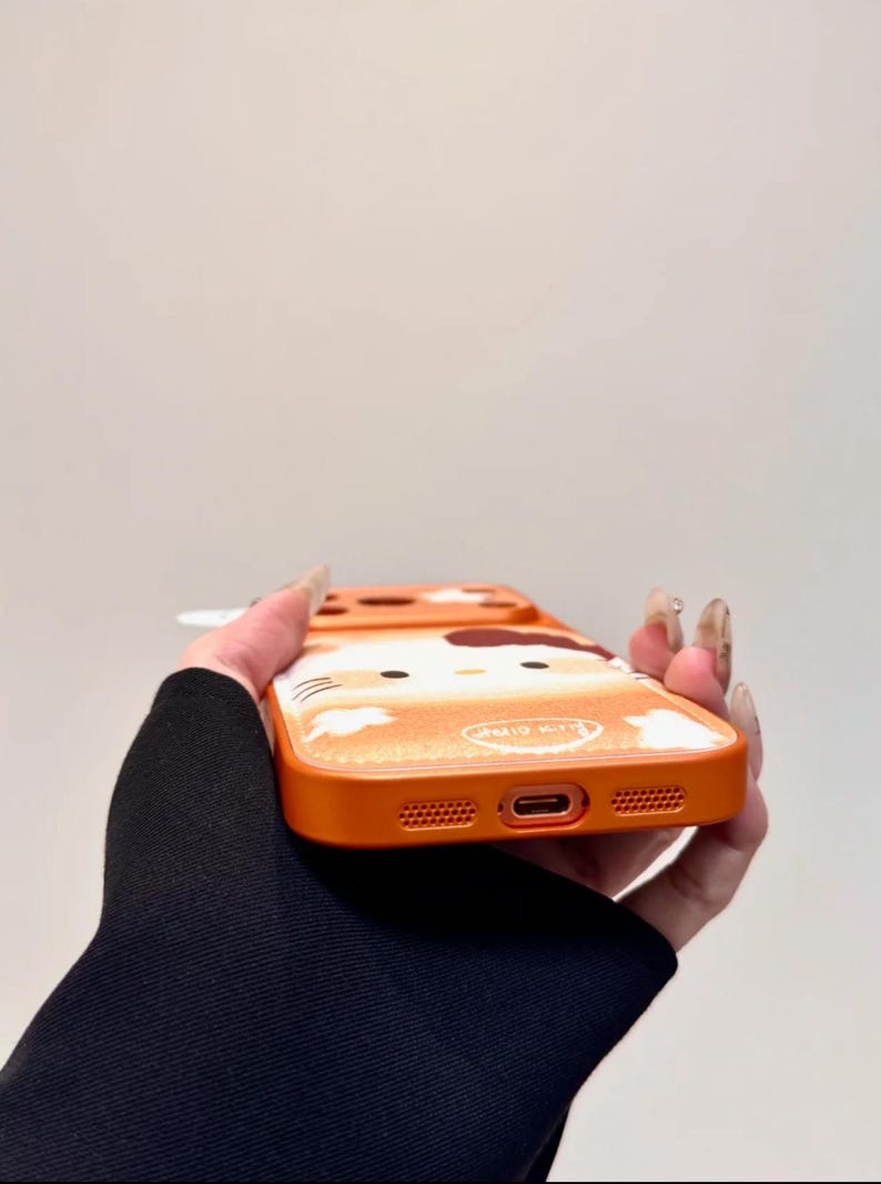 May include: Orange phone case with a Hello Kitty design. The case has a glittery finish and a cartoon image of Hello Kitty. It has cutouts for the phone's camera, charging port, and speakers. The text "Hello Kitty" is visible.
