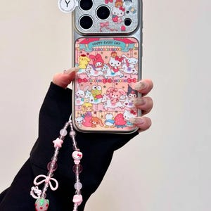 K&ouml;nnte beinhalten: Silberne Handyh&uuml;lle mit einem Karussell-Design mit Zeichentrickfiguren und dem Text "HAPPY EVERY DAY". Sie hat einen passenden Perlenriemen mit rosa Perlen, Sternen und einem Erdbeer-Anh&auml;nger.
