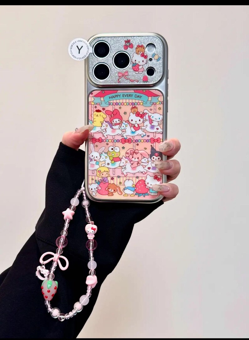 K&ouml;nnte beinhalten: Silberne Handyh&uuml;lle mit einem Karussell-Design mit Zeichentrickfiguren und dem Text "HAPPY EVERY DAY". Sie hat einen passenden Perlenriemen mit rosa Perlen, Sternen und einem Erdbeer-Anh&auml;nger.