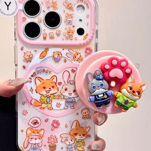 May include: Clear phone case with a pink border and a cartoon animal design. The case features a heart-shaped graphic with a fox and bunny, plus a pop-up grip with animal figures and a paw print.