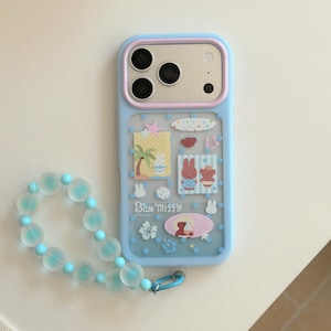 May include: A light blue smartphone case with a pink camera frame and a translucent back featuring Miffy character stickers, blue polka dots, and the text Blue Miffy. A matching wrist strap made of frosted light blue beads is attached to the case.